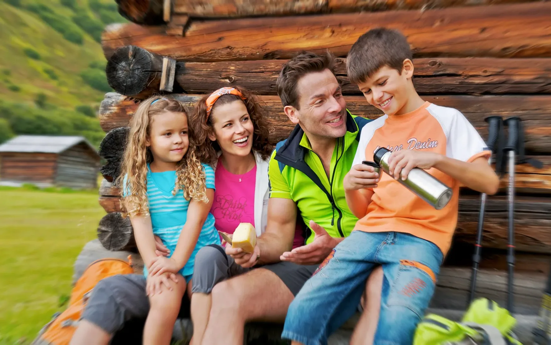 Eine Gruppe von Menschen, darunter zwei Erwachsene und zwei Kinder, sitzt draußen auf einer Holzkonstruktion. Ein Kind reicht einem anderen eine Wasserflasche, während alle lächeln. Im Hintergrund ist Wanderausrüstung zu sehen.