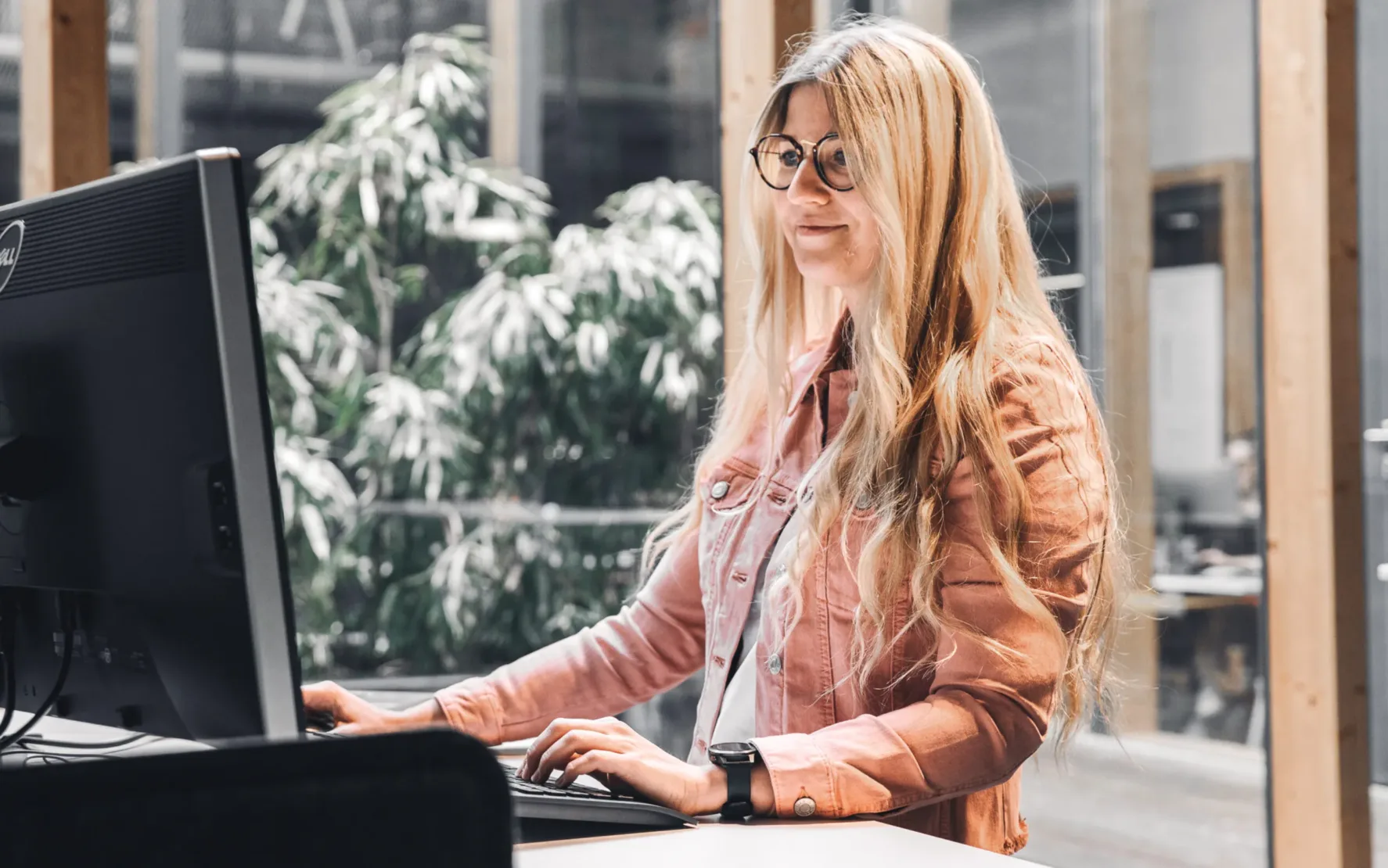 Bild von einer jungen Frau mit Brille und blonden Haaren, die an einem Steharbeitsplatz am Computer arbeitet.