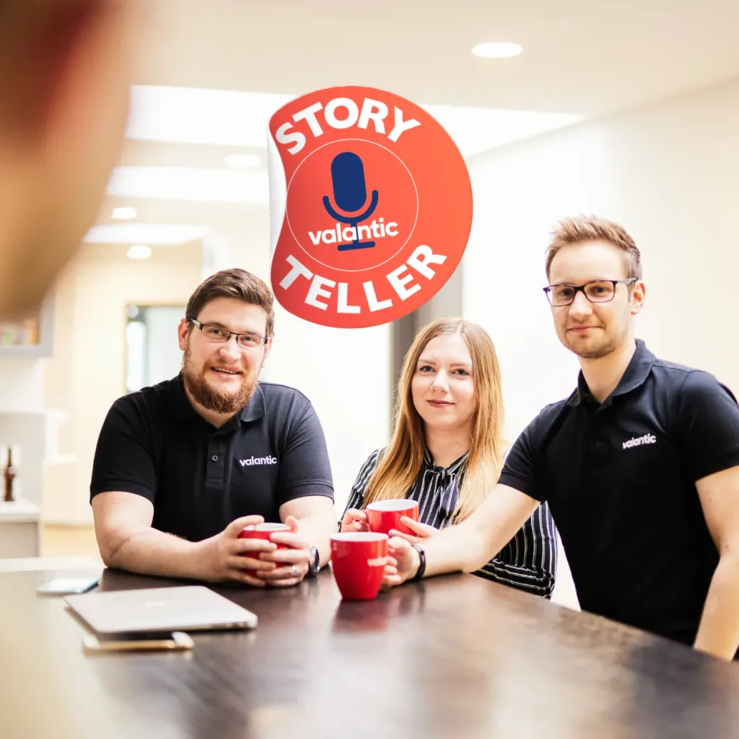 Drei Personen in einem modernen Büro stehen neben einem Holztresen und halten rote Tassen in der Hand. Sie tragen schwarze Hemden mit „Valantic“-Logos. Im Hintergrund ist ein rotes, rundes „Story Teller“-Logo zu sehen.