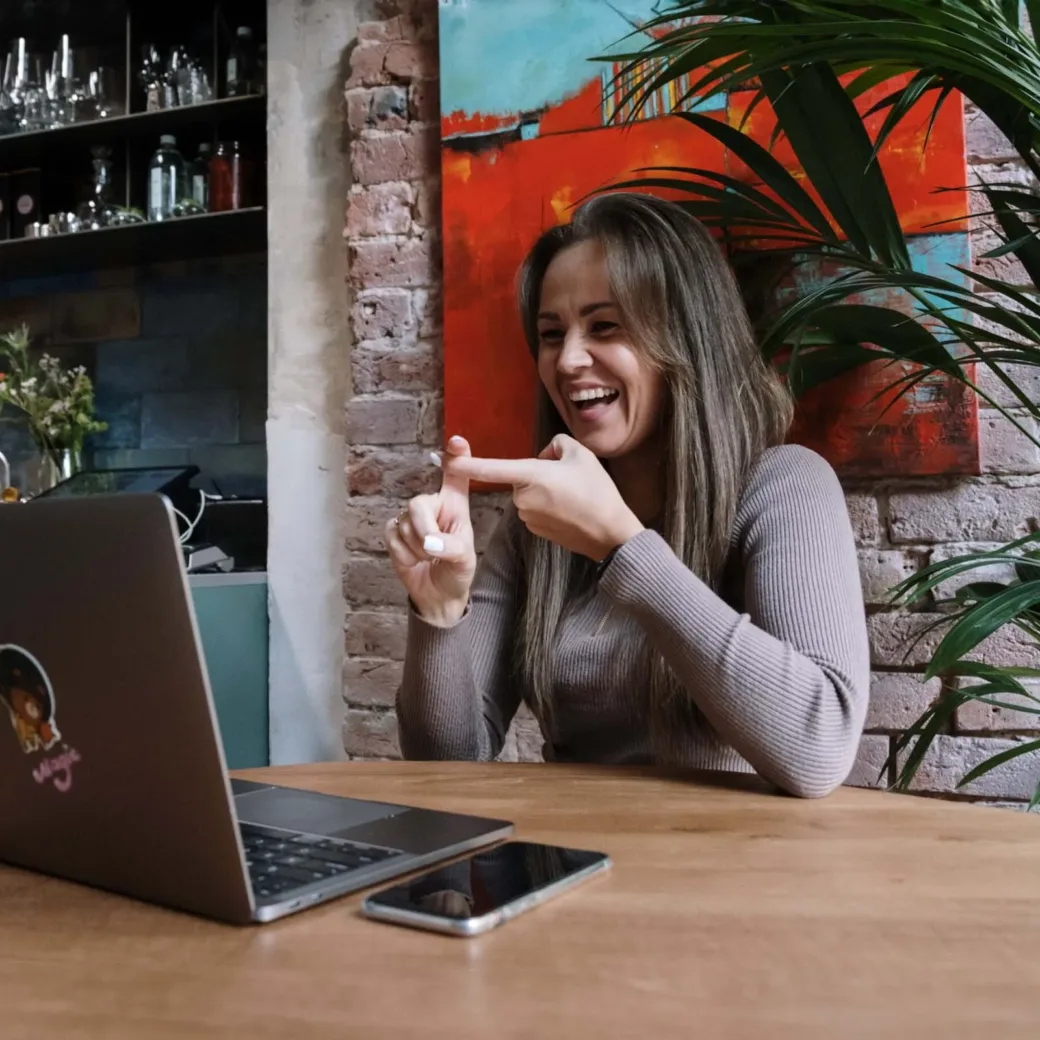 A woman with long hair, wearing a light brown sweater, uses sign language at a wooden table with a laptop and smartphone in a modern kitchen setting.